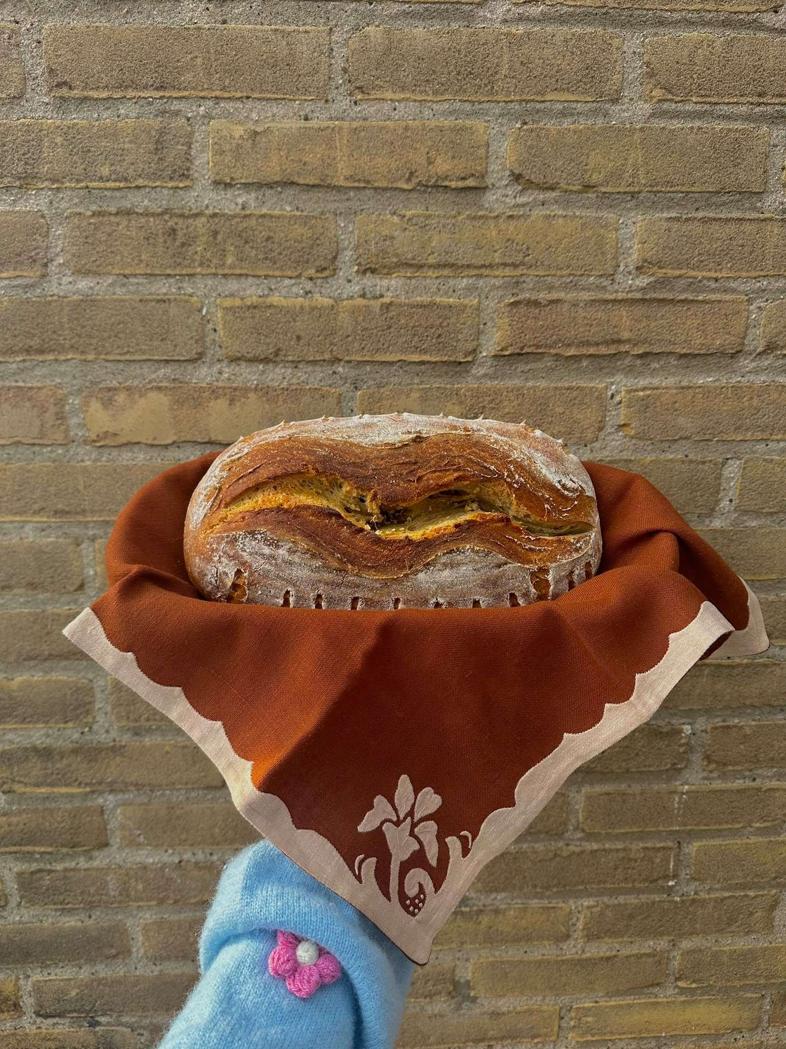 Een vers gebakken zuurdesembrood met een diepe insnijding, gewikkeld in een roestbruine linnen doek voor een gele bakstenen muur